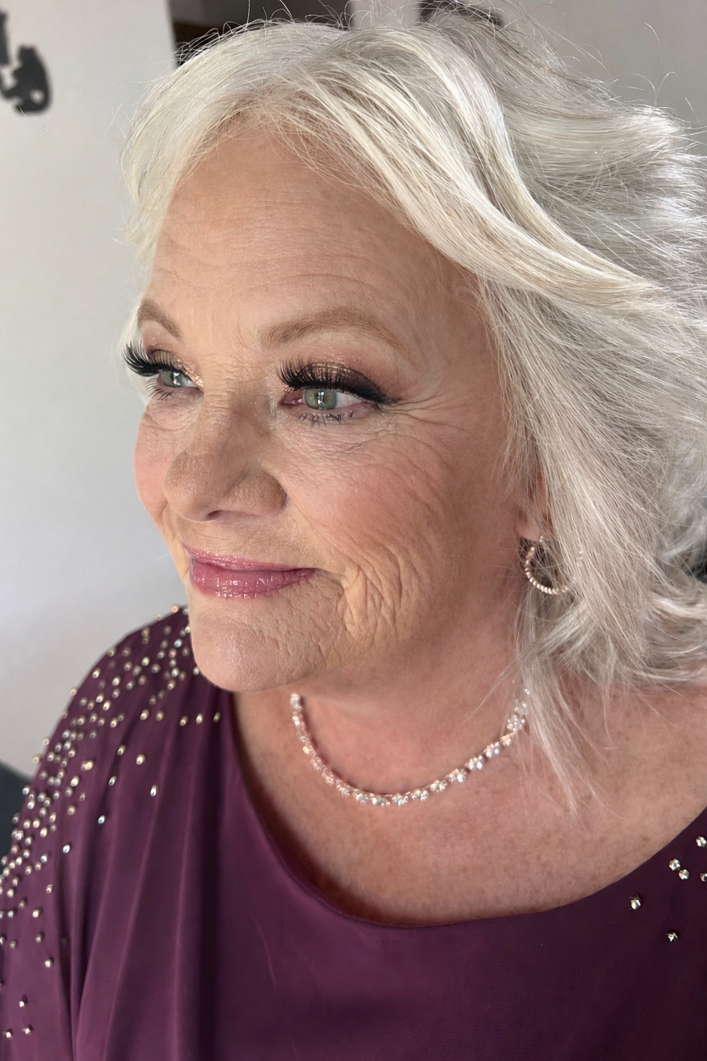 Close-up of an elderly woman with silver hair, wearing a purple dress with pearl embellishments, a pearl necklace, and earrings, smiling.