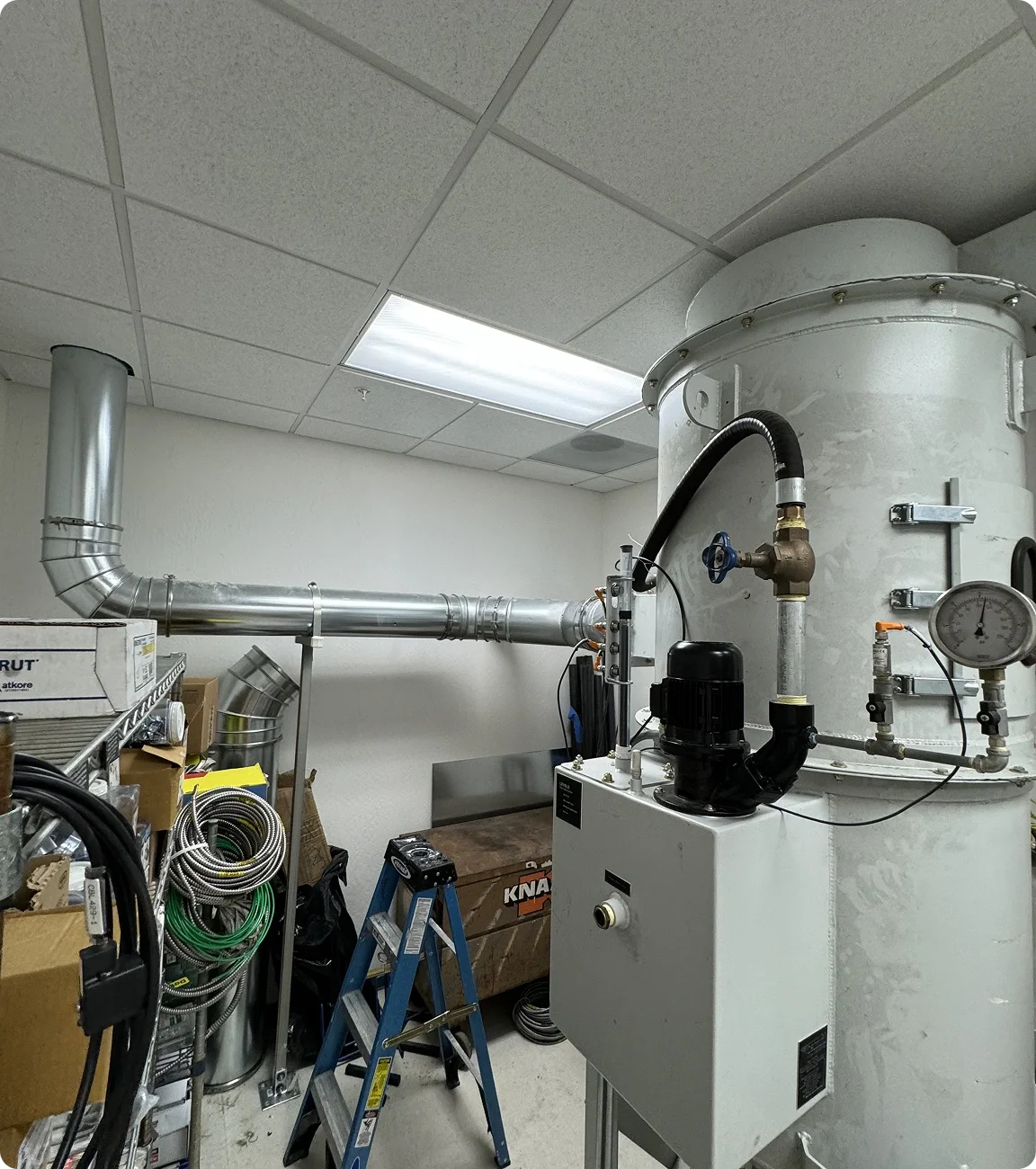 Industrial machinery and pipes in a utility or mechanical room with a ladder and storage boxes.