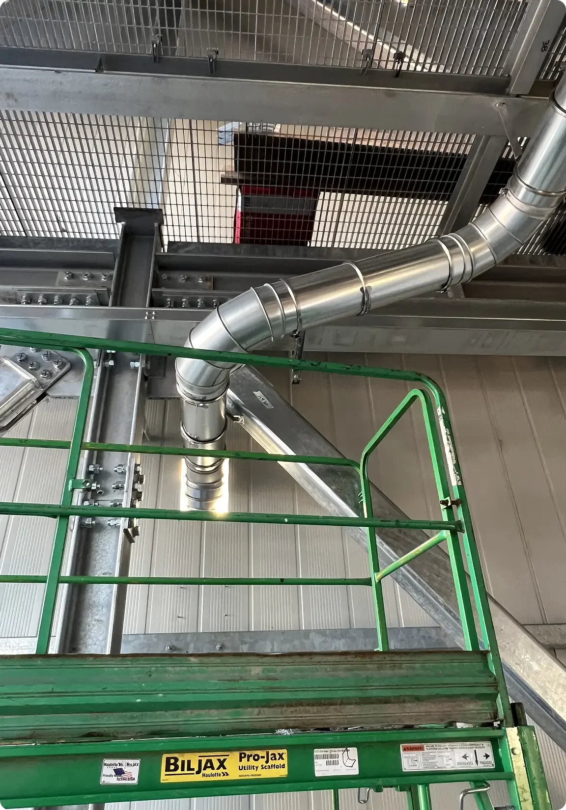 View from a green man lift showing a metal ventilation duct, a metal grate, and part of a ceiling in an industrial or construction setting.