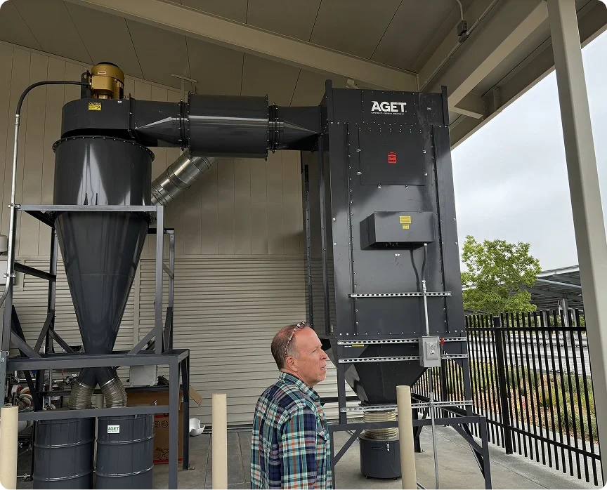 A man standing in front of an industrial dust collection system outdoors, with a fence and trees in the background.