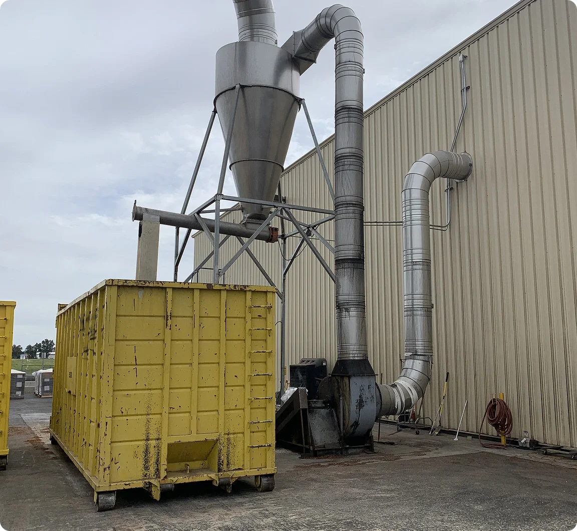 Industrial dust collection system with large metal pipes and a yellow dumpster in front, set against a beige metal building and cloudy sky.