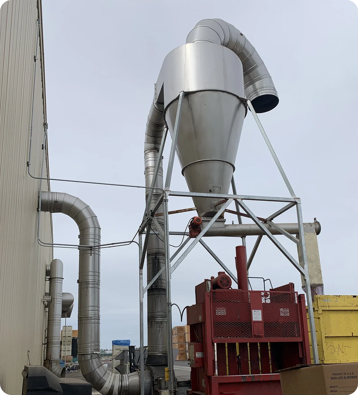 Industrial dust collection system with large metal pipes connected to a collection hopper, situated outside a building with a red machine and storage containers.
