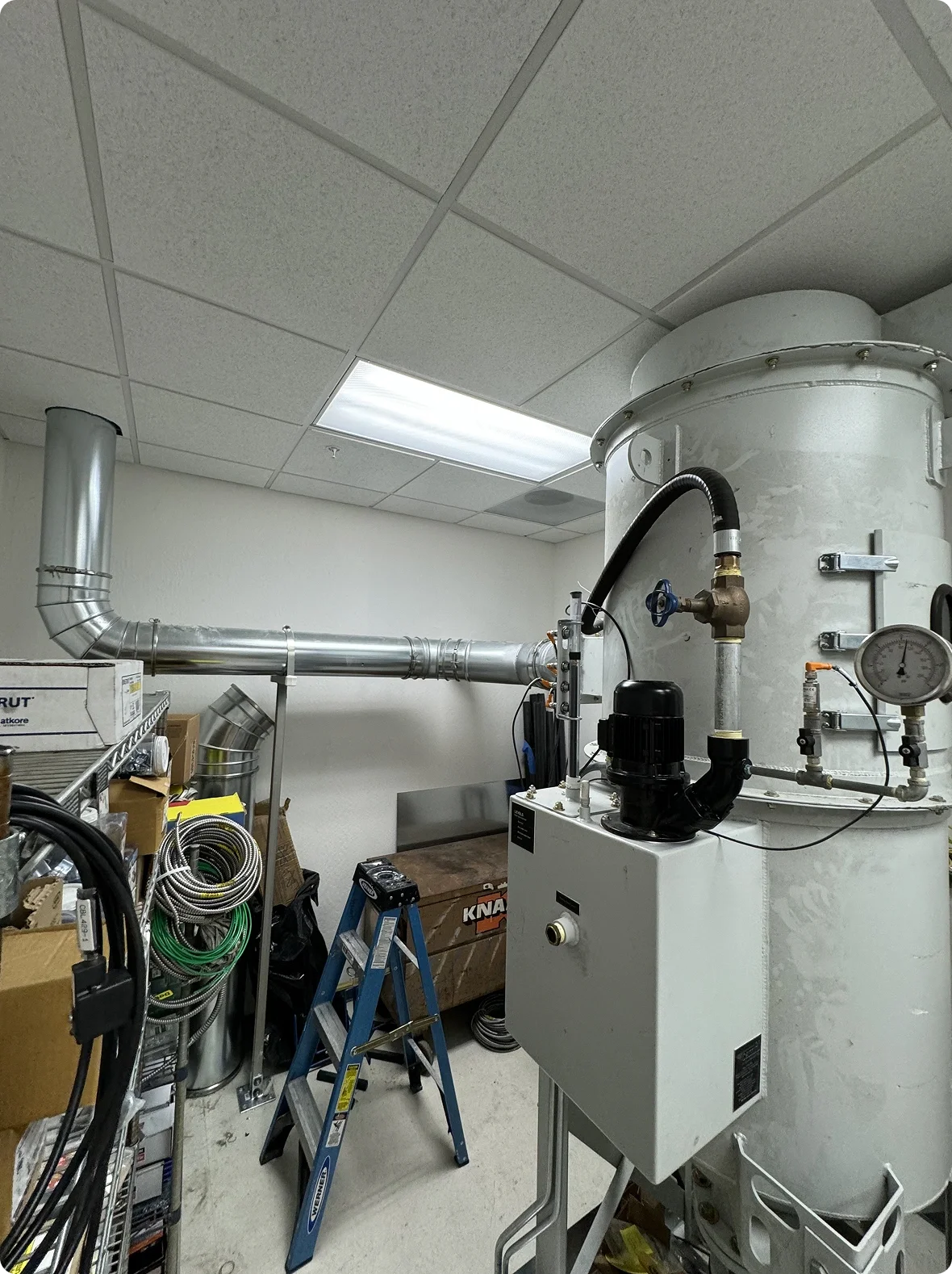 Industrial mechanical equipment including a large vertical cylindrical tank, pipes, gauges, and tools in a room with a drop ceiling.