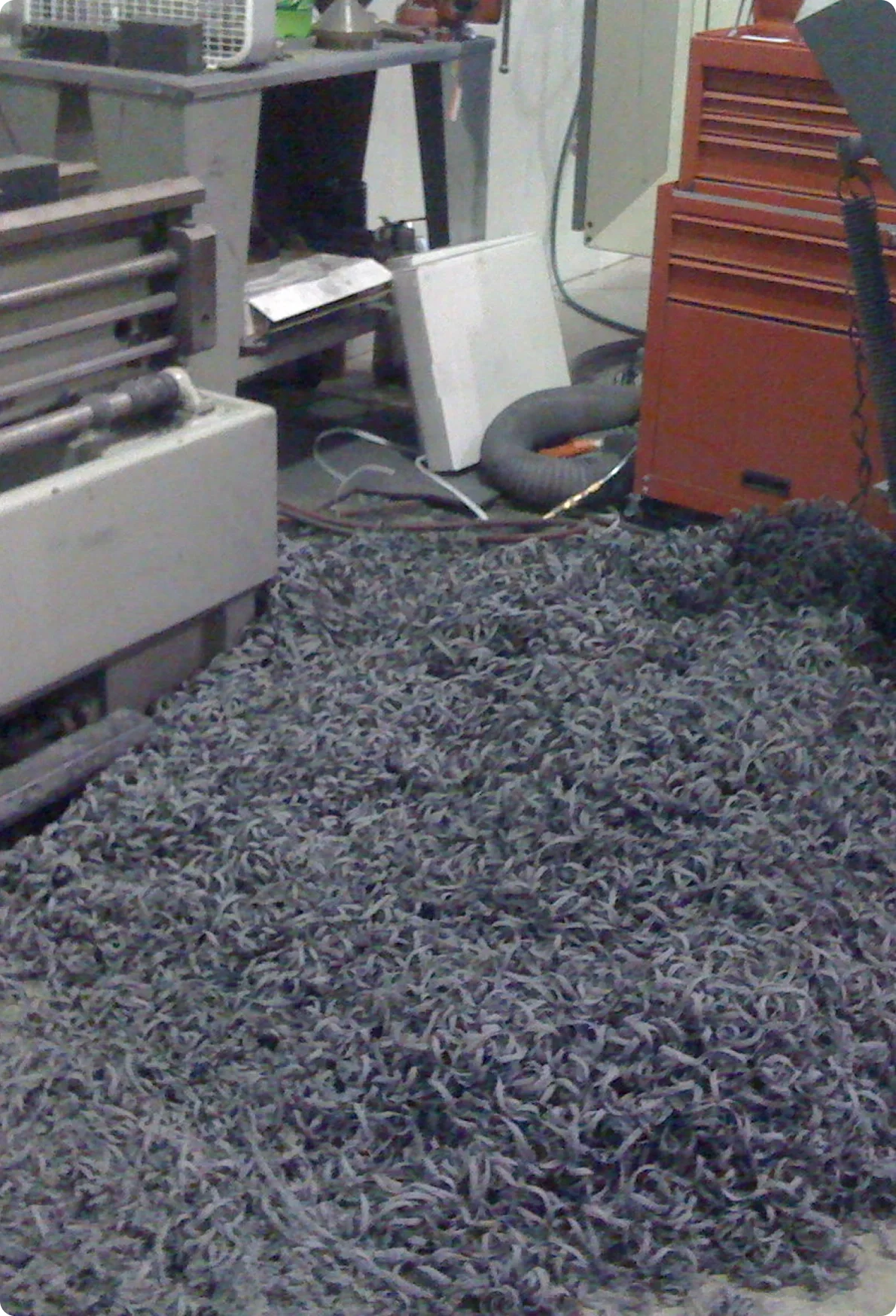 A workshop or garage space with tools, equipment, and a cluttered workbench. A vacuum hose is on the floor near a white panel and a carpeted rug covers the floor.