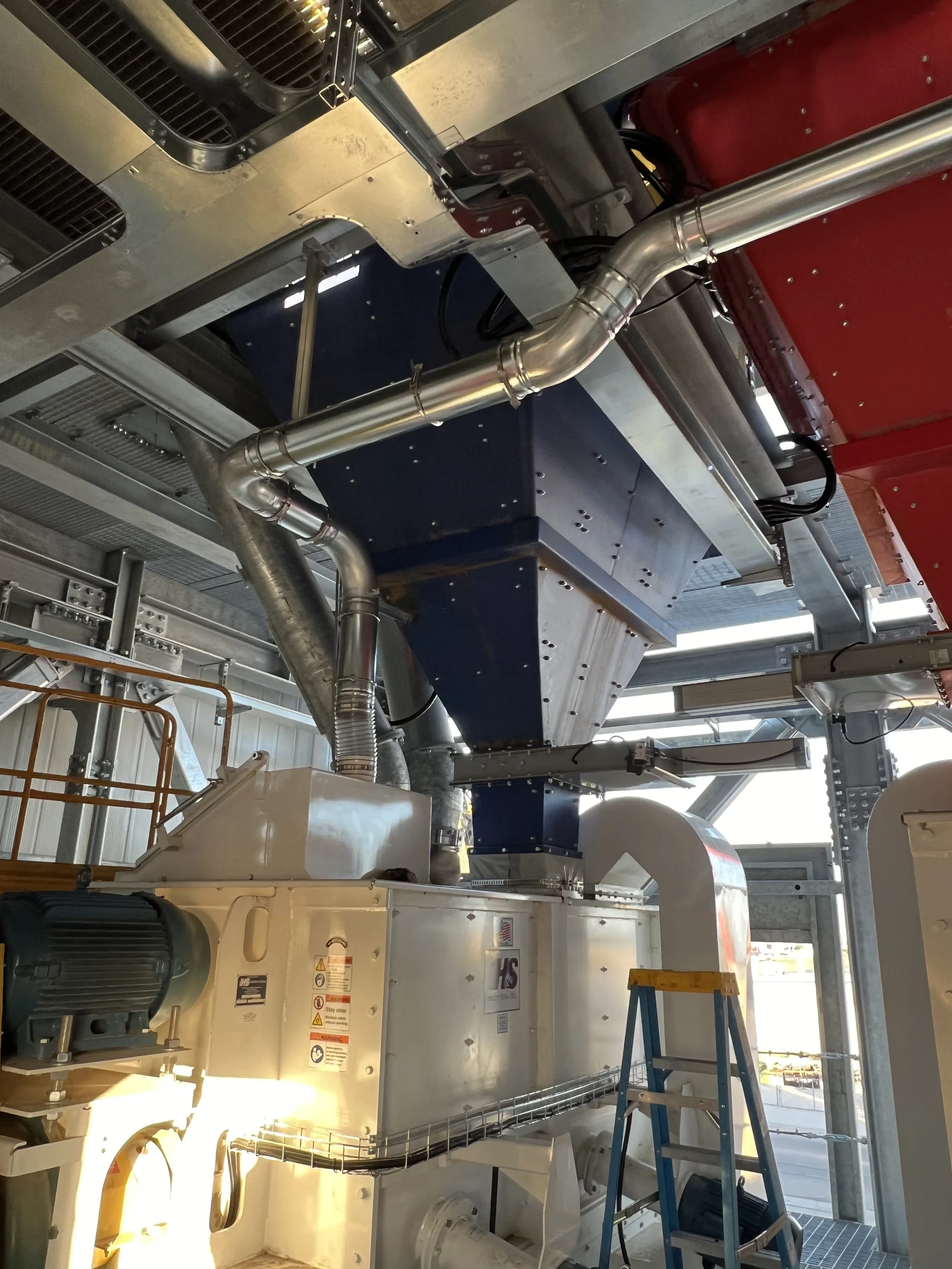 Industrial machinery inside a building with metal pipes, ducts, and a ladder.