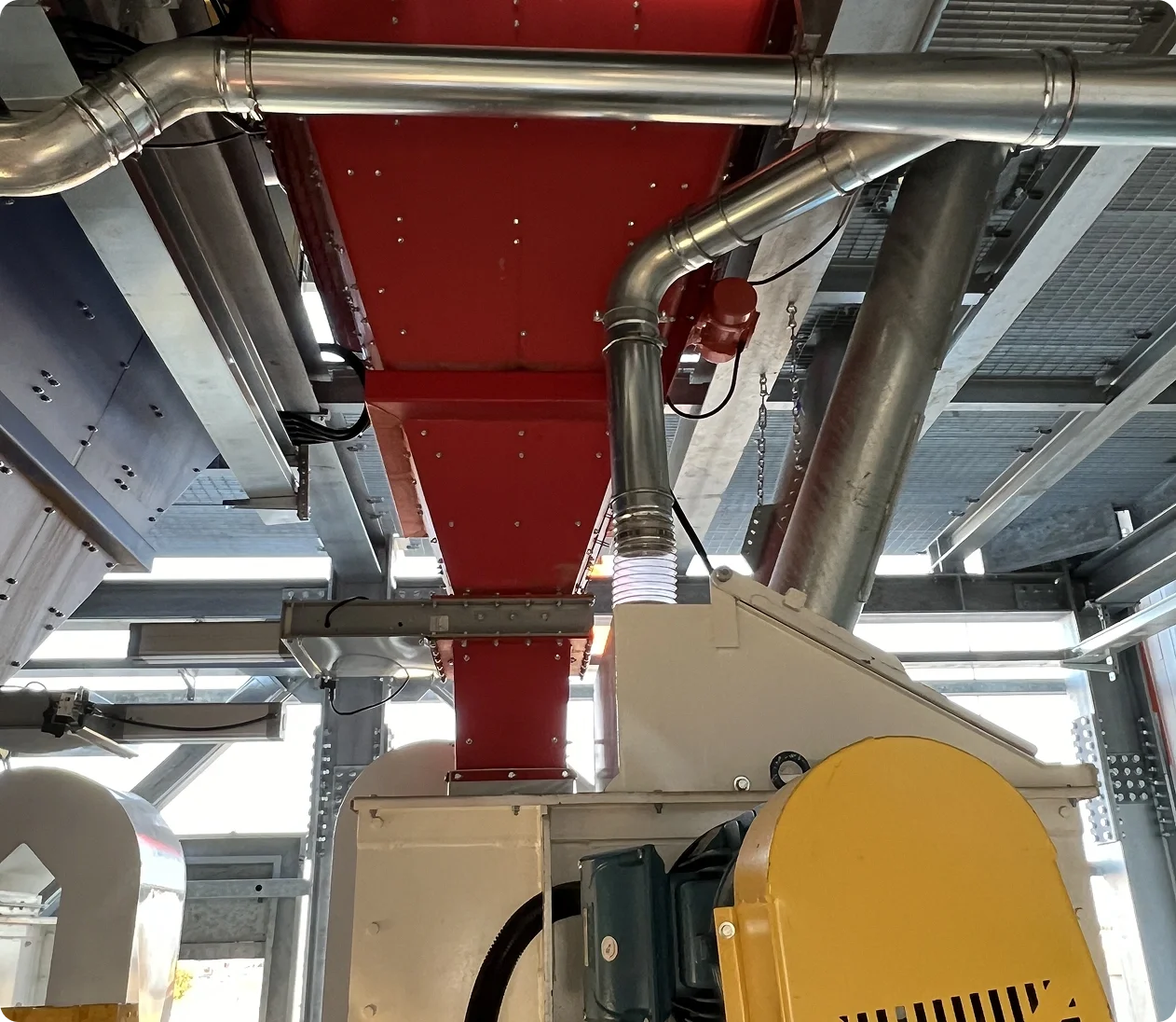 Image of industrial machinery with metallic pipes and ducts, including a red component, a large metallic pipe, and a yellow device, situated within a steel framework.