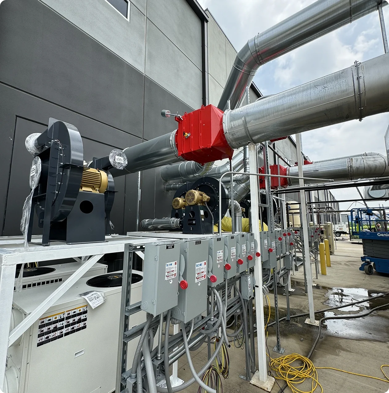 Industrial piping system with metal pipes, valves, and electrical equipment outside a building under a cloudy sky.