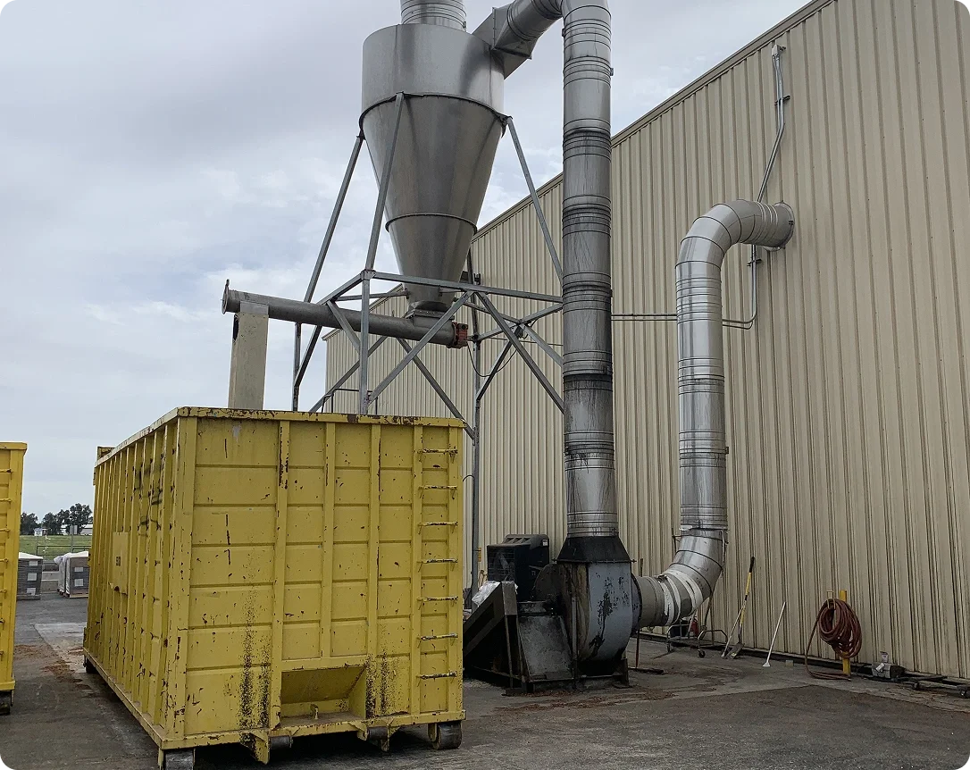 Industrial ventilation system with two large metal exhaust pipes connected to a beige building, a yellow dumpster in the foreground, and some tools and hoses along the wall.