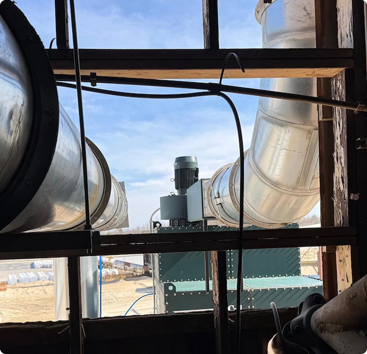 View of industrial ducts and ventilation equipment seen through a wooden and metal framework, with a clear blue sky in the background.