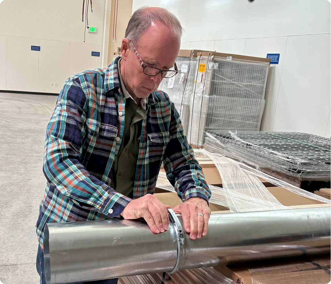 A man in plaid shirt and glasses working with a large metal barrel or pipe in an industrial or warehouse environment.