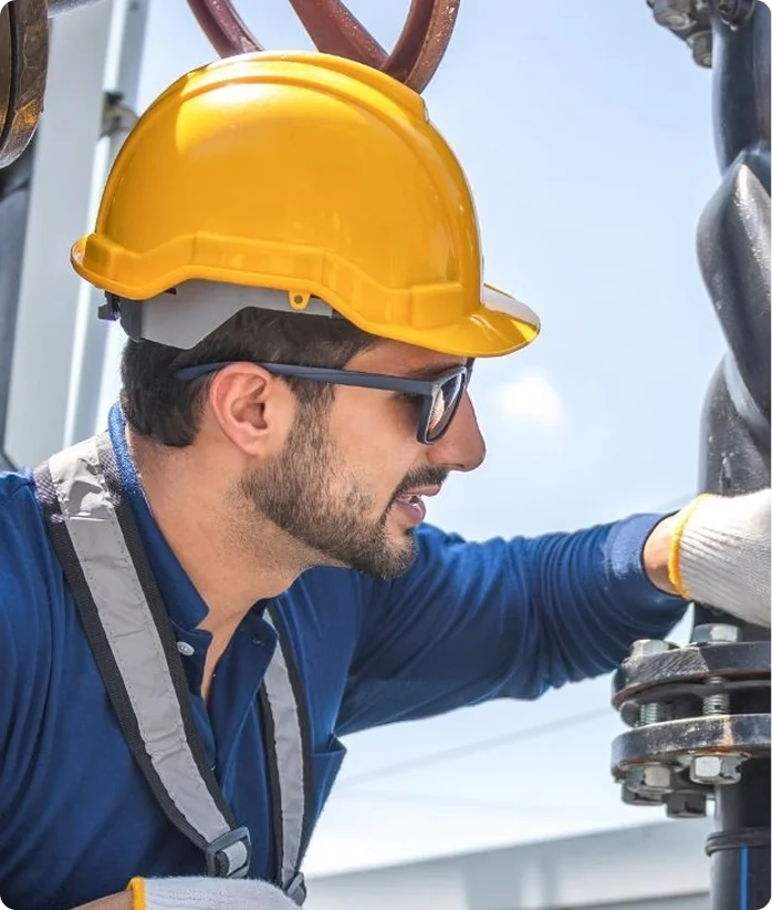 A man wearing sunglasses and a yellow safety helmet working outdoors on a mechanical or electrical task.