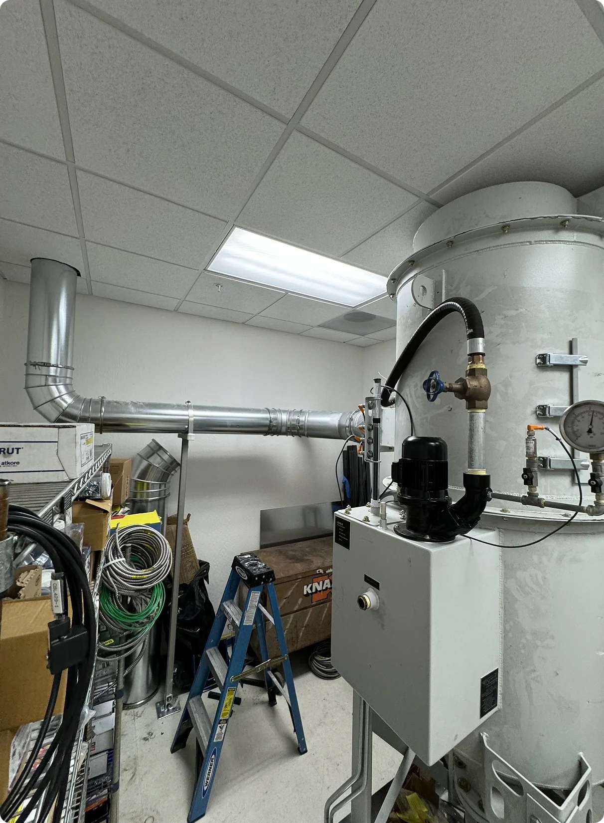 A mechanical room with pipes, gauges, and equipment, including a large vertical white tank and various ducts and hoses.