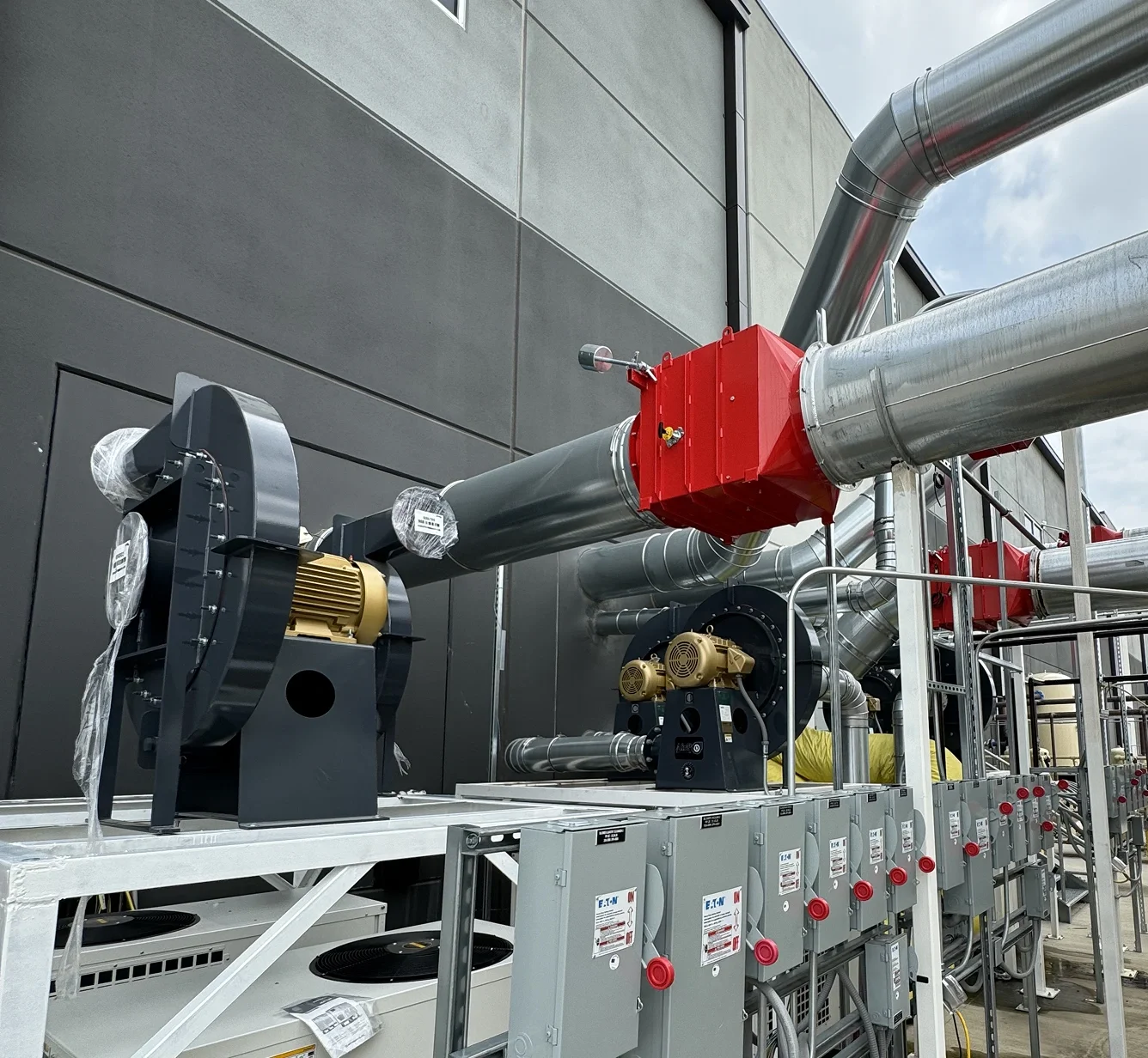 Industrial piping system with metallic pipes, red junction boxes, and electrical control panels mounted on a gray support structure outside a building.