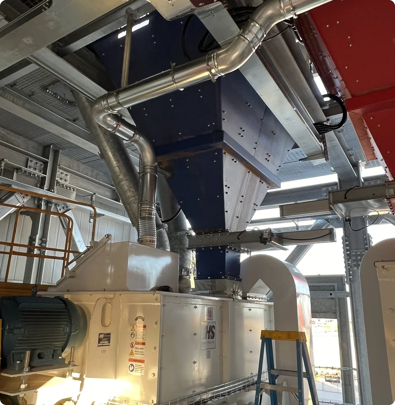 Industrial machinery with metal ducts and a large white control unit, inside a building with metal framework and a ladder.