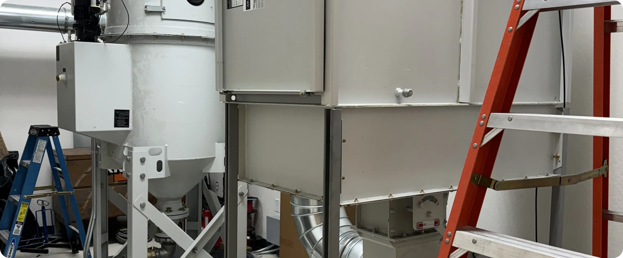 Industrial equipment including a large white machine, orange ladder, and various pipes and control boxes in a workshop or utility room.