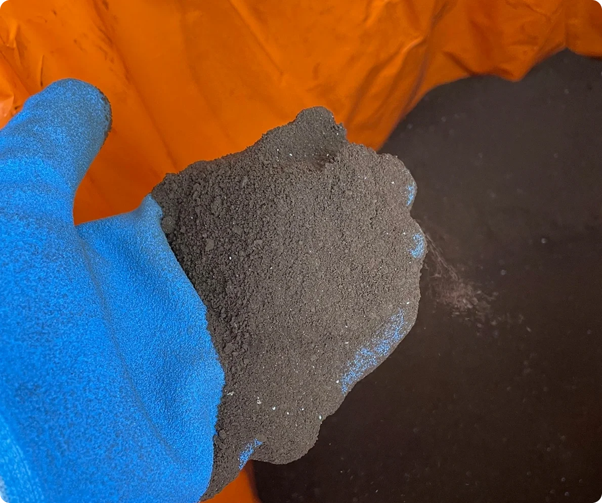 A gloved hand holding a clump of black granular material, with an orange background and black granules on the surface below.