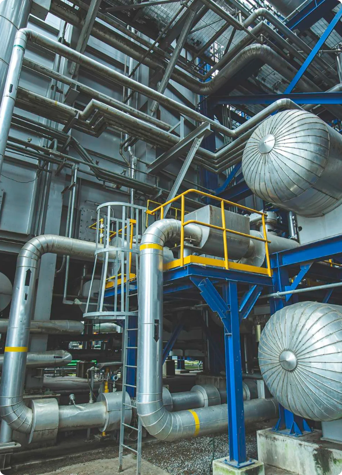 Industrial piping and machinery with large silver pipes, yellow safety railings, and blue structural supports.