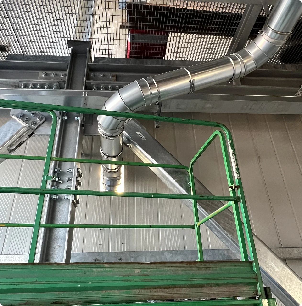 View from a platform showing a metal industrial pipe running diagonally across the image, with a green railing in the foreground, and part of a metal mesh barrier at the top.