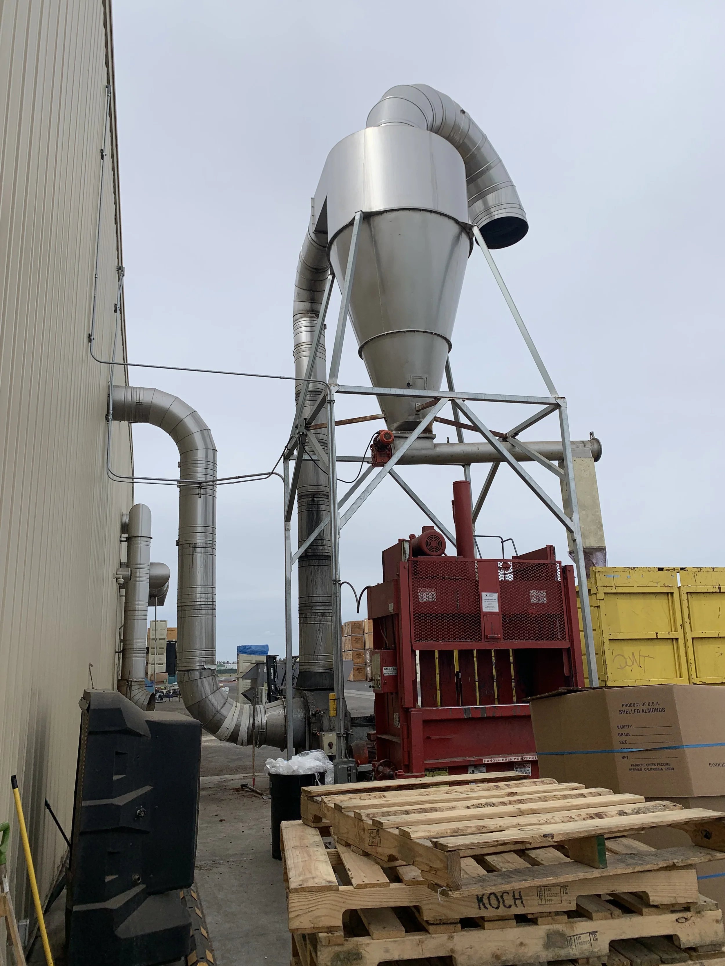 Industrial outdoor scene with large metal dust collector with duct pipes, wooden pallets, and a yellow container.