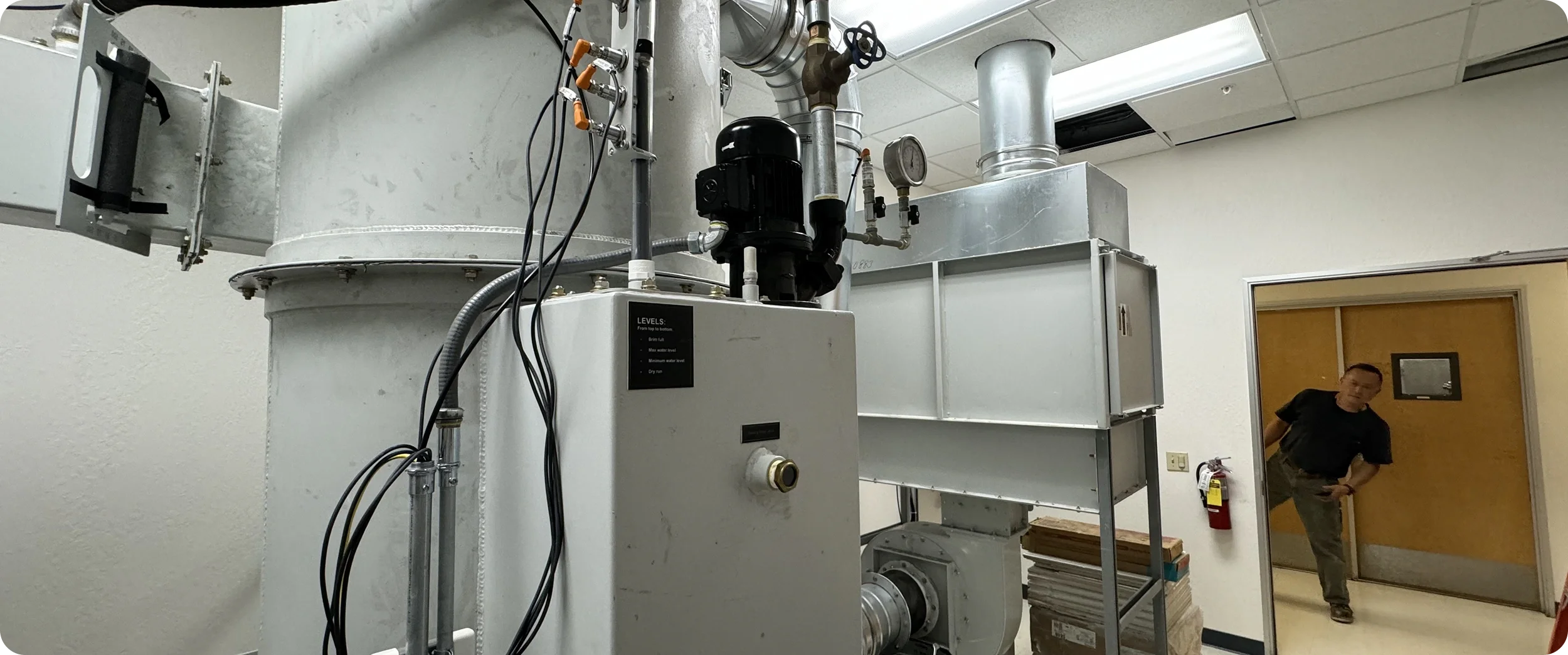 Industrial machinery with pipes, gauges, and electrical wiring in a room, with a man standing at a doorway in the background.