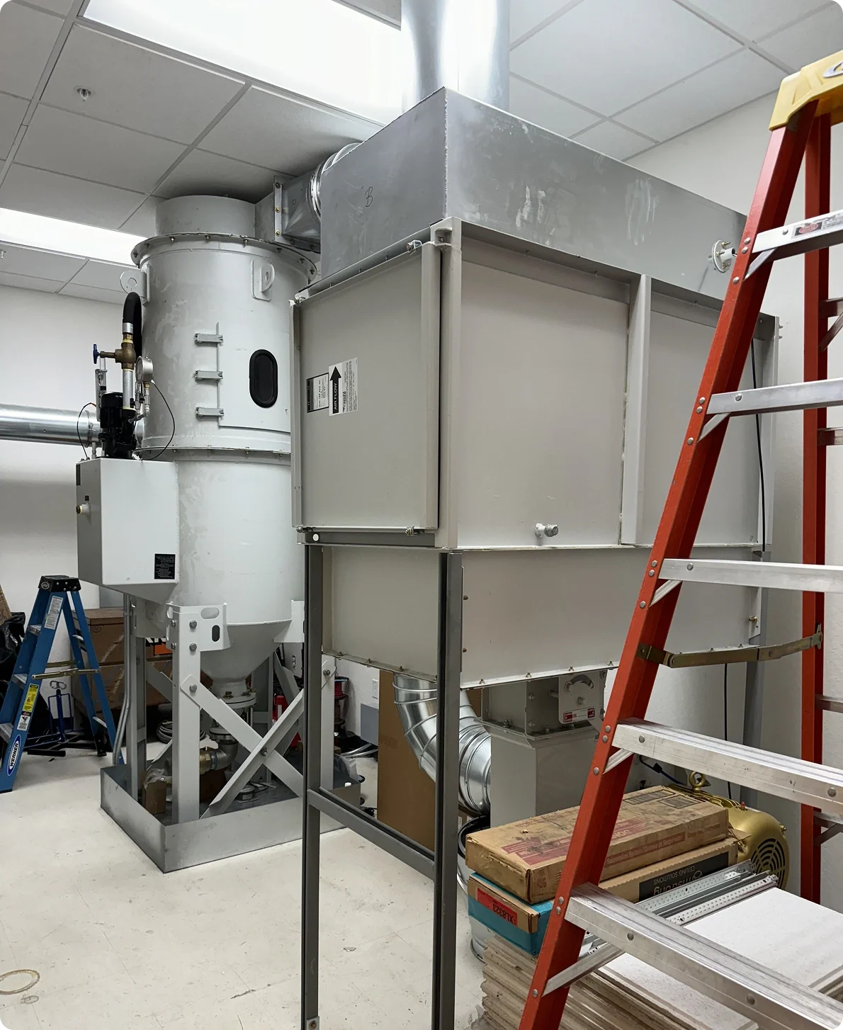 Industrial machine with ductwork and electrical components in a utility or manufacturing room, with a stepladder and various tools nearby.