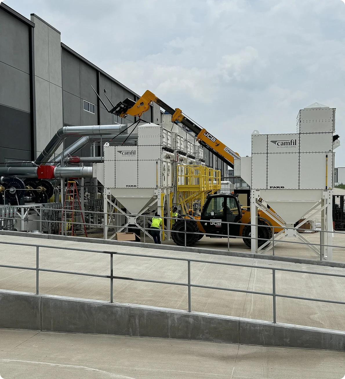 Industrial scene with large white equipment labeled 'Camfil' for air pollution control, a JCB crane lifting a component, and workers in safety vests nearby.
