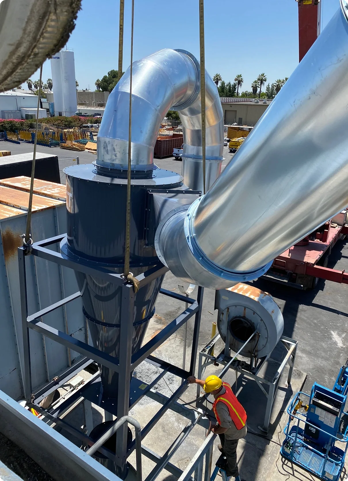 Large industrial ventilation or duct system under construction with shiny metallic pipes, a worker in a yellow helmet and orange safety vest working nearby, and a clear blue sky in the background.