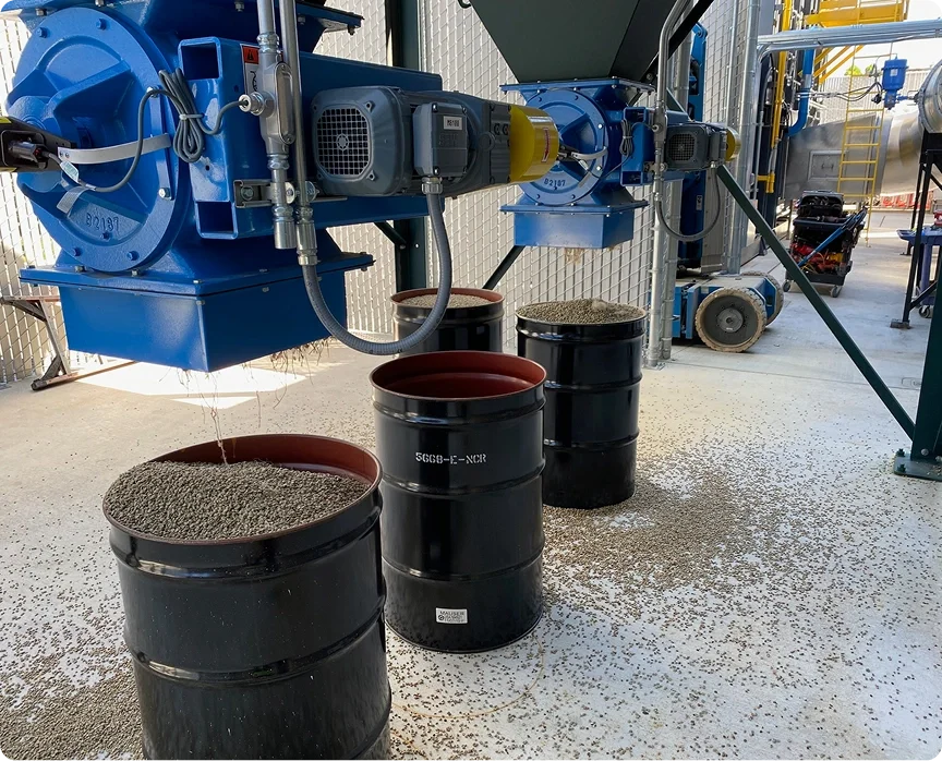 Industrial machinery processing or manufacturing using barrels filled with granular material, possibly in a factory or processing plant.