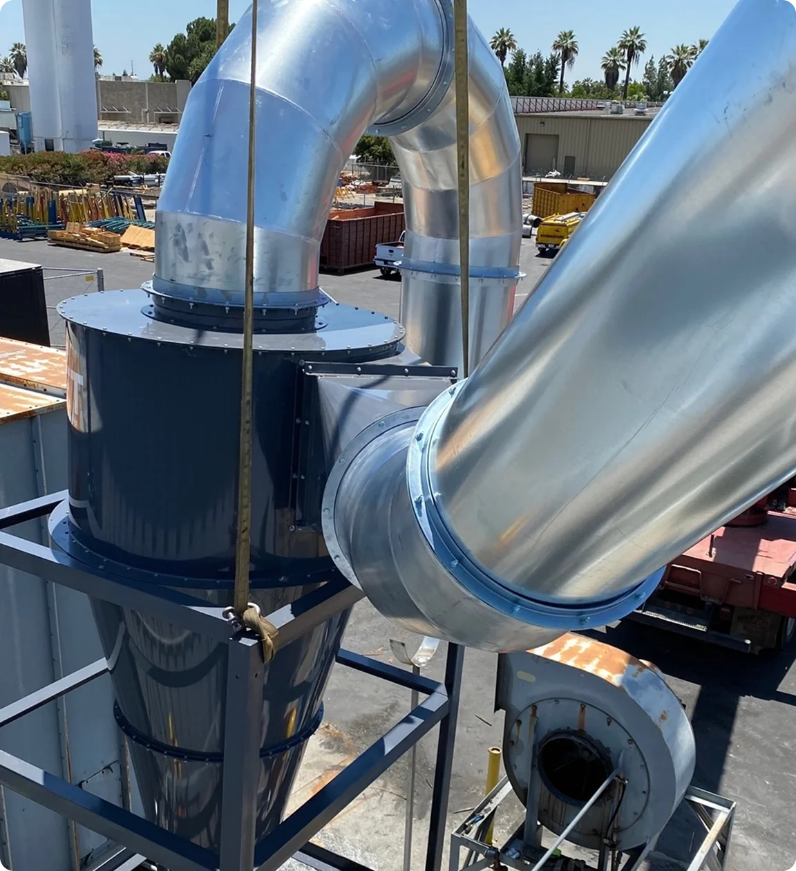 Large industrial metal ducts and pipes in an outdoor industrial setting with palm trees in the background.