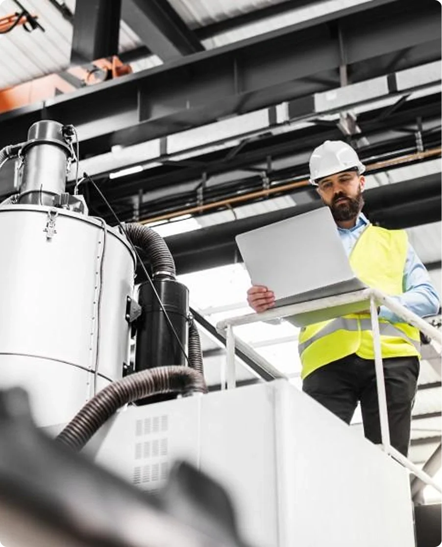 A man wearing a white hard hat and yellow safety vest is standing on a platform, looking at a laptop inside an industrial facility with machinery and pipes overhead.