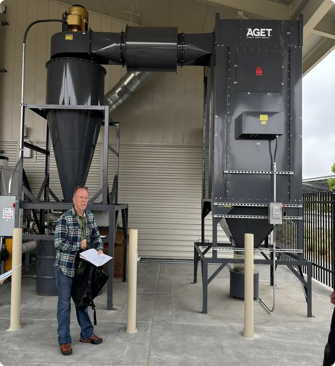 Large industrial air filtration or dust collection system with ducts and a control panel, outside a building, with a man holding papers standing nearby.