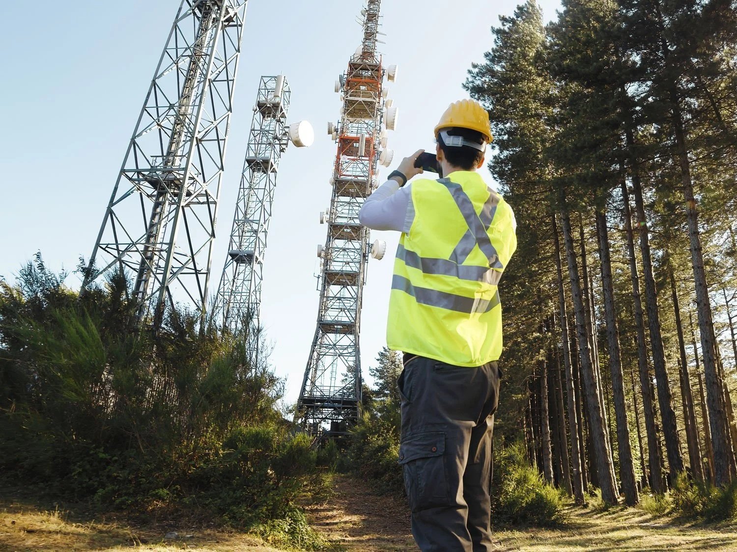 A worker wearing a yellow safety vest and hard hat takes a photo of radio towers in a wooded area during daylight.