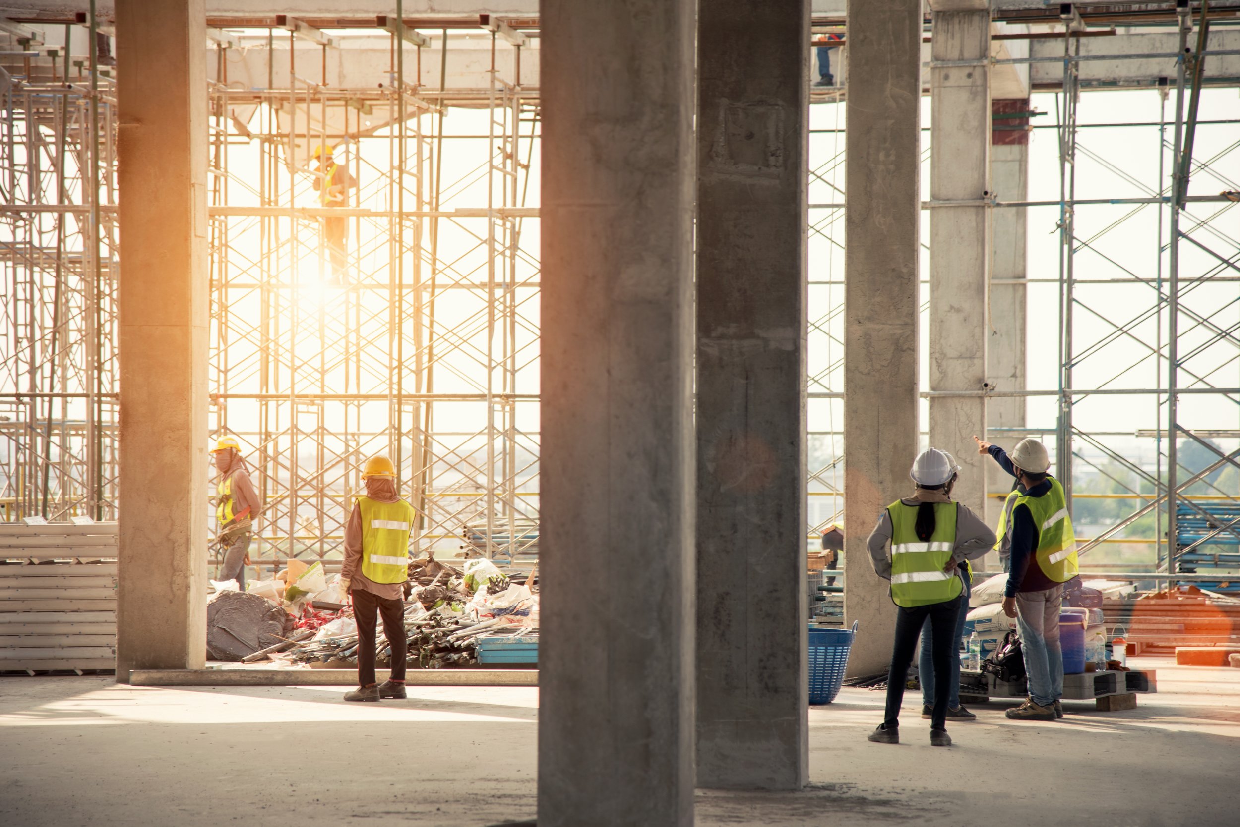Construction workers wearing safety vests and helmets on a building site with scaffolding, exposed concrete columns, and sunlight in the background.