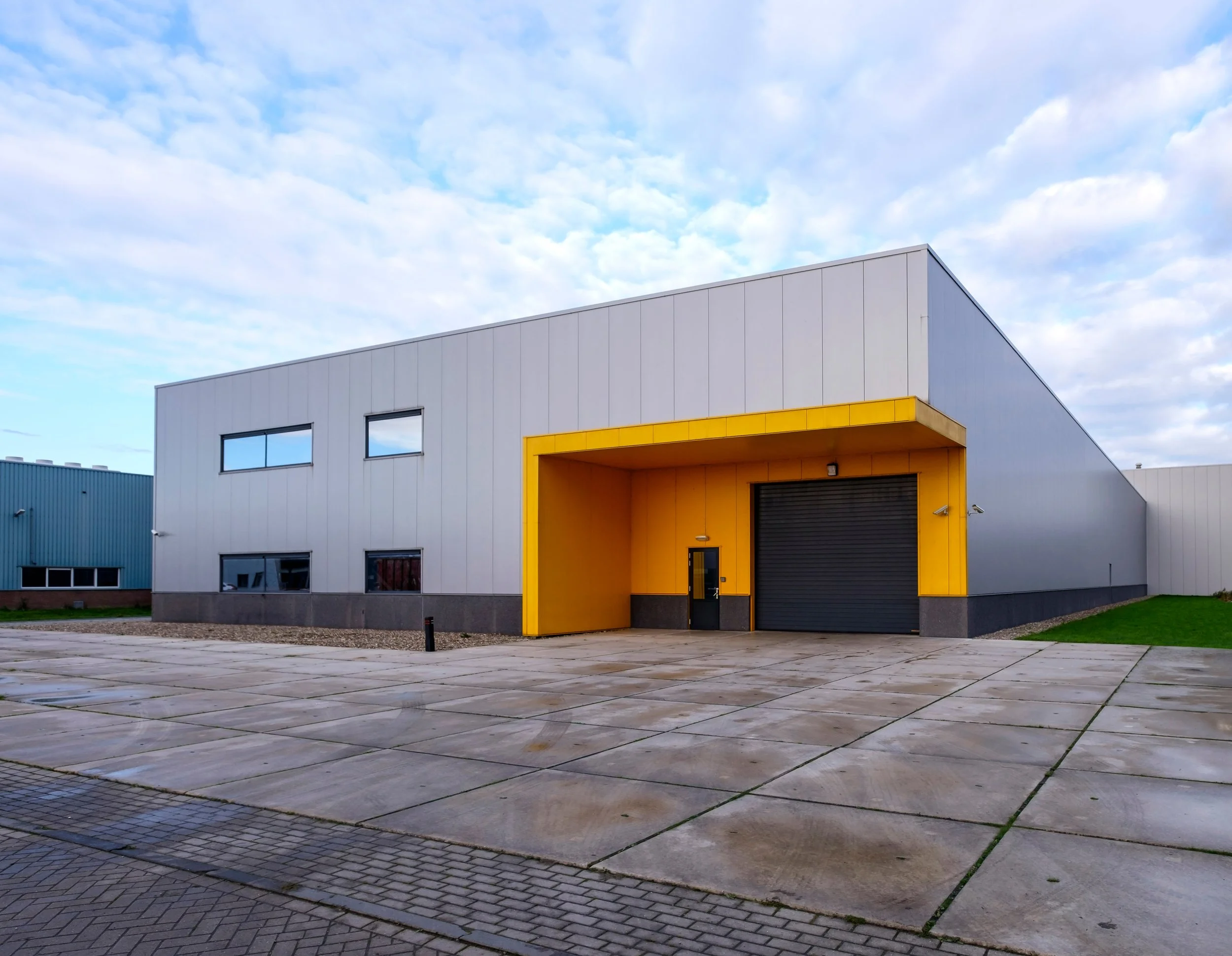 Modern industrial building with white and gray exterior walls, accented by a prominent yellow entryway. The building is situated on a paved lot with a grid pattern, and the sky above is partly cloudy with patches of blue.