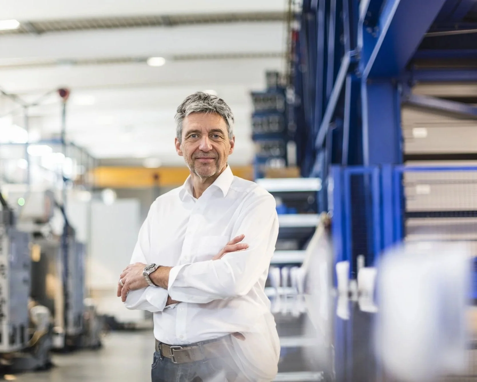 A man in a white shirt and grey trousers standing with arms crossed in an industrial or manufacturing setting with blue equipment and workstations behind him.