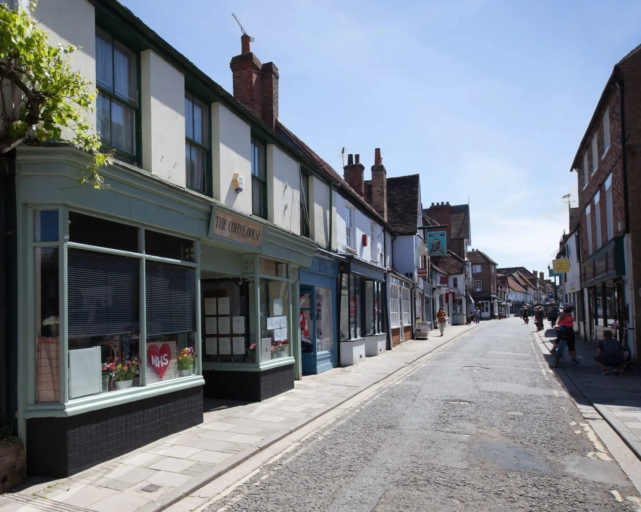 A row of shops on a sunny street with people walking. The storefronts include a coffee shop with a large window display and a sign reading 'THE COFFEE HOUSE'. The buildings are multi-story, with some having brick chimneys and antennas on the roofs. There are trees along the sidewalk.