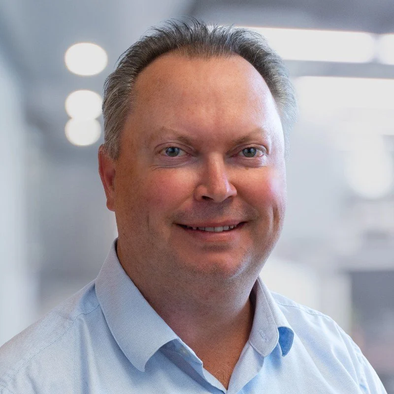 Portrait of John Wood wearing a light blue shirt, in an indoor office setting with blurred background lighting.