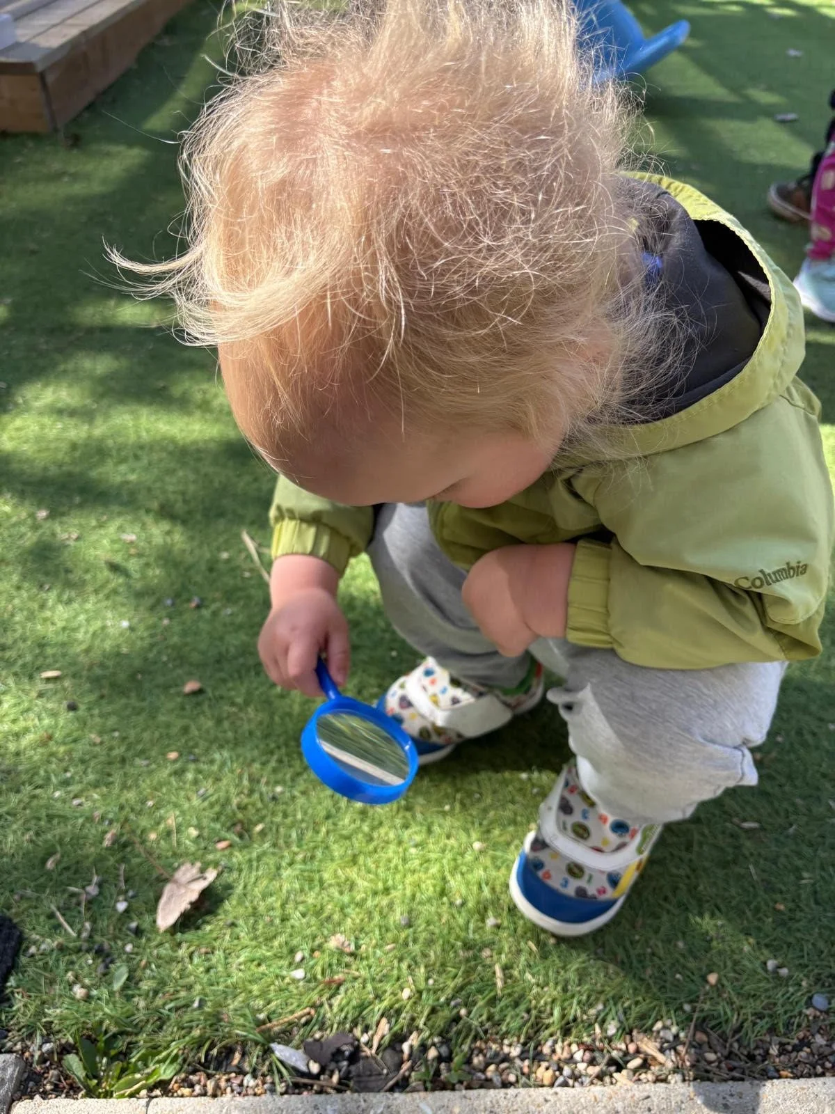 A toddler examining the grass outside using a blue toy magnifying glass during a daycare activity.