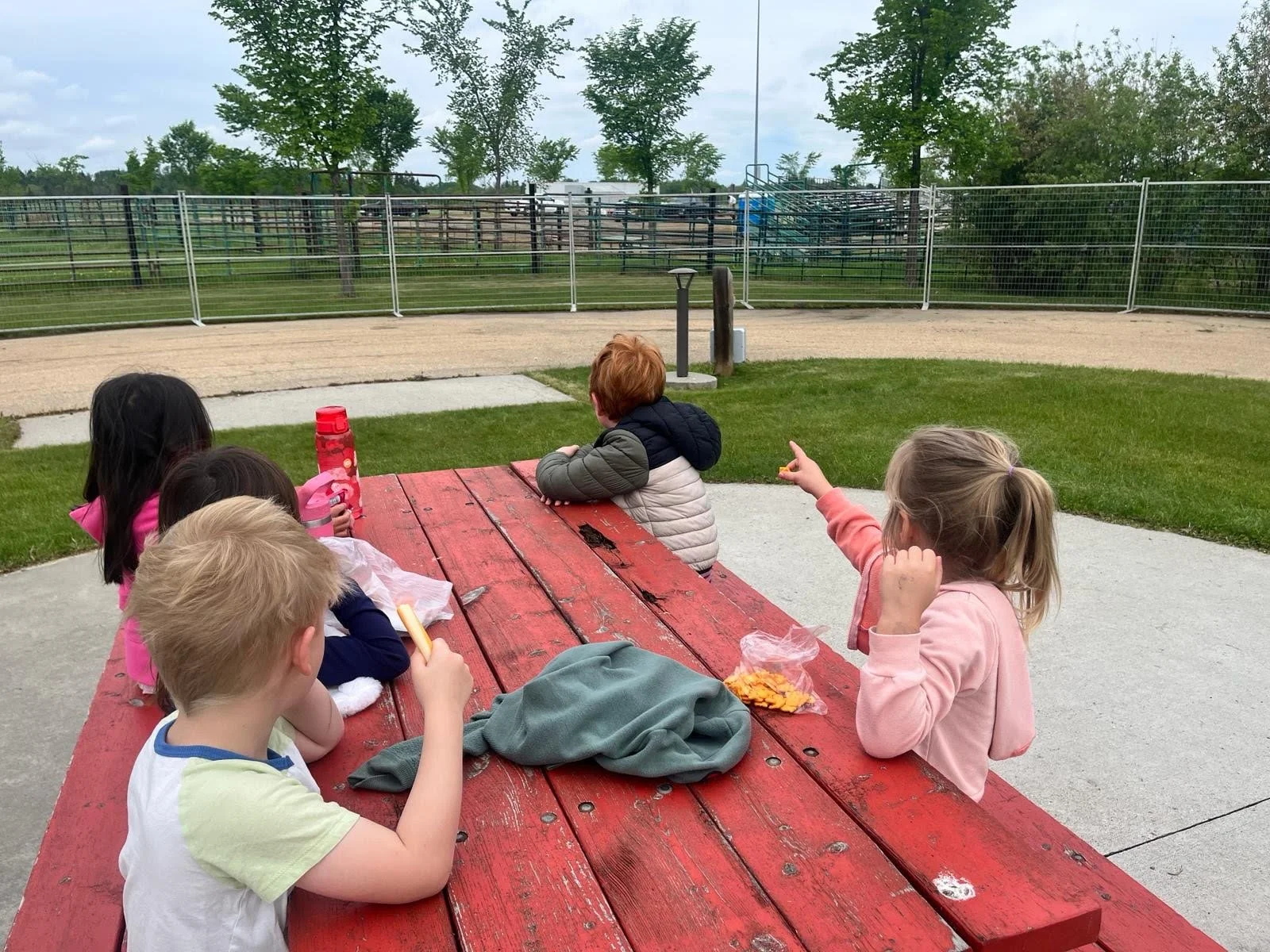 A group of young children sitting together at a red wooden picnic table outside while out exploring the community.