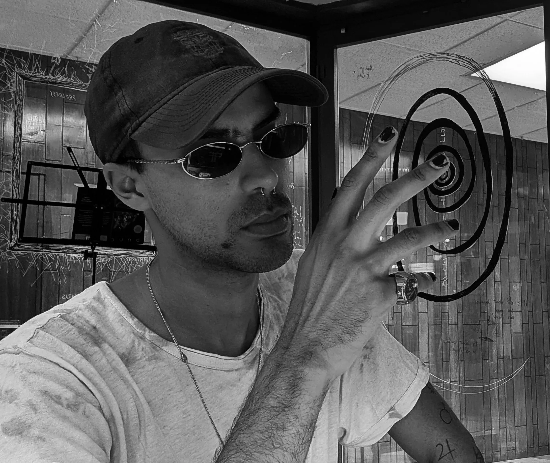 A person wearing sunglasses, a hat, and a nose ring is taking a selfie in front of a glass window with a spiral designed on it. The person is pointing at the spiral with their finger.