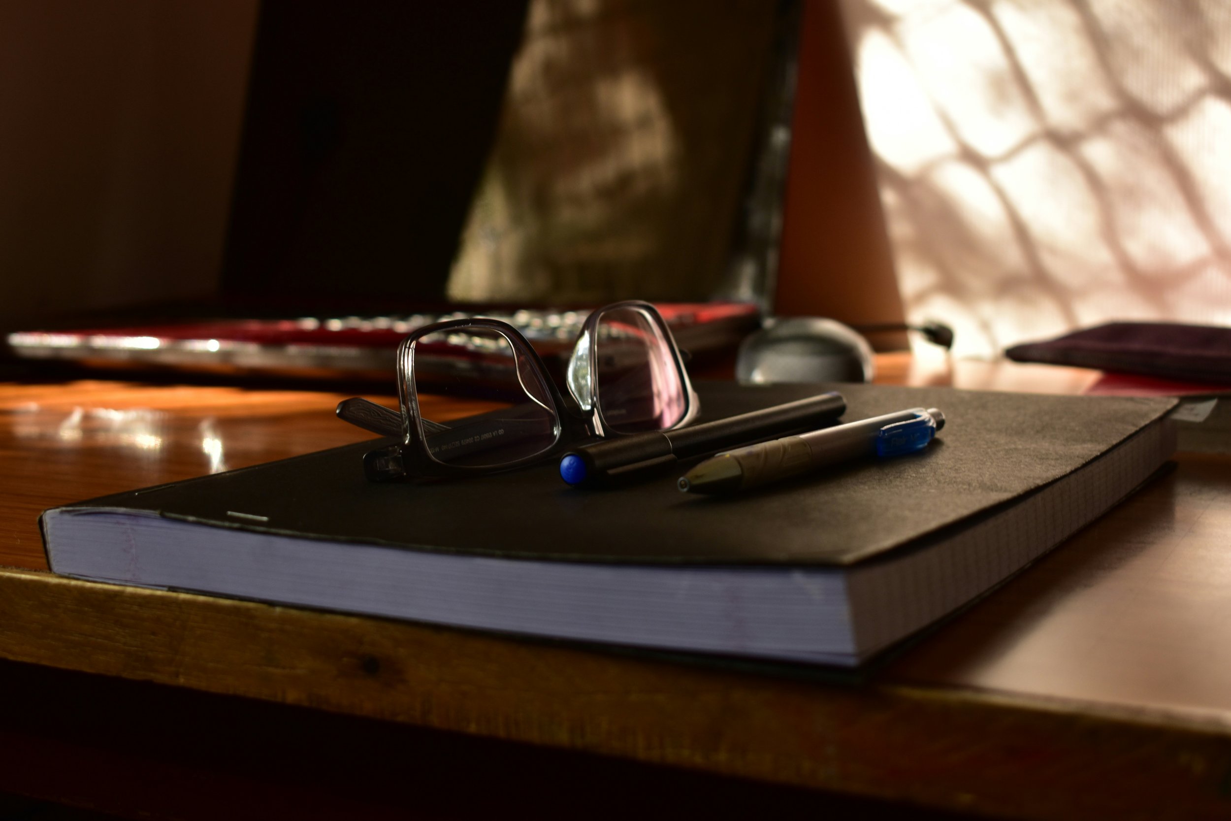 A pair of glasses, a black pen, a yellow highlighter, and a blue pen resting on a closed black notebook on a wooden desk.