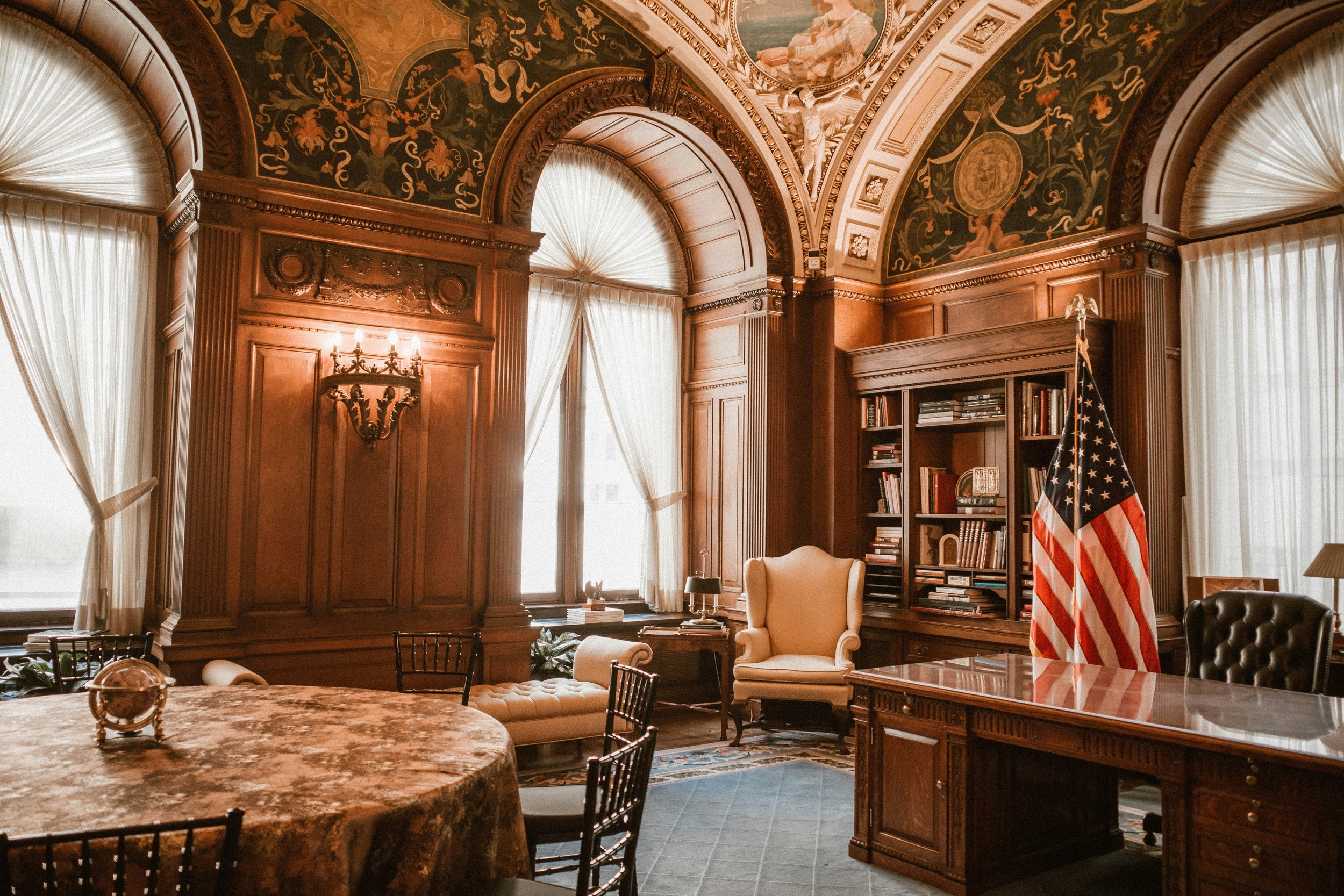 A room with wood-paneled walls and ornate ceiling artwork, large windows with curtains, a bookshelf with books, an American flag, a cream-colored armchair, and a marble-topped table with a decorative globe.
