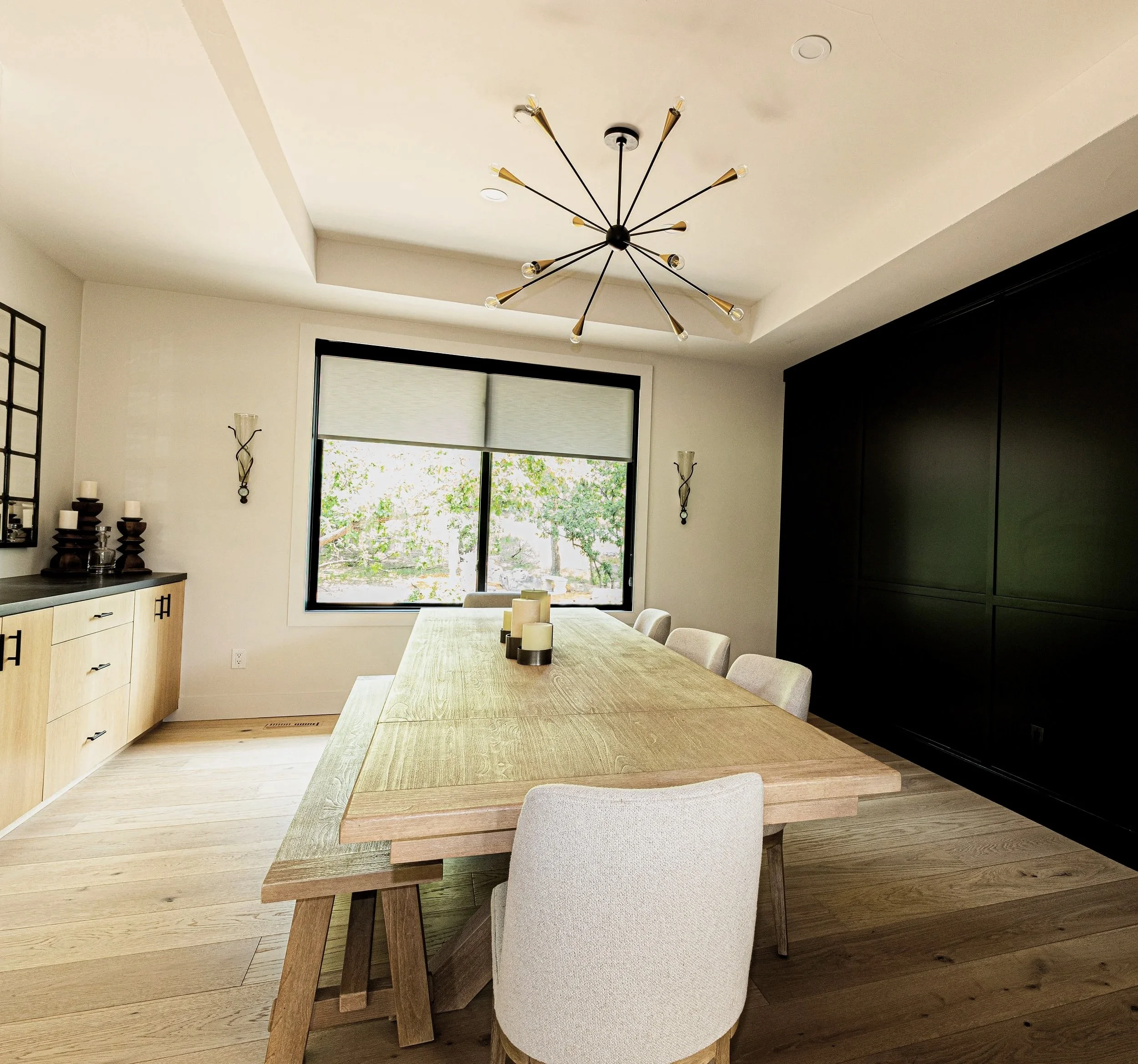 A modern dining room with a wooden table, beige upholstered chairs, a black cabinet, and a large window with a view of trees.