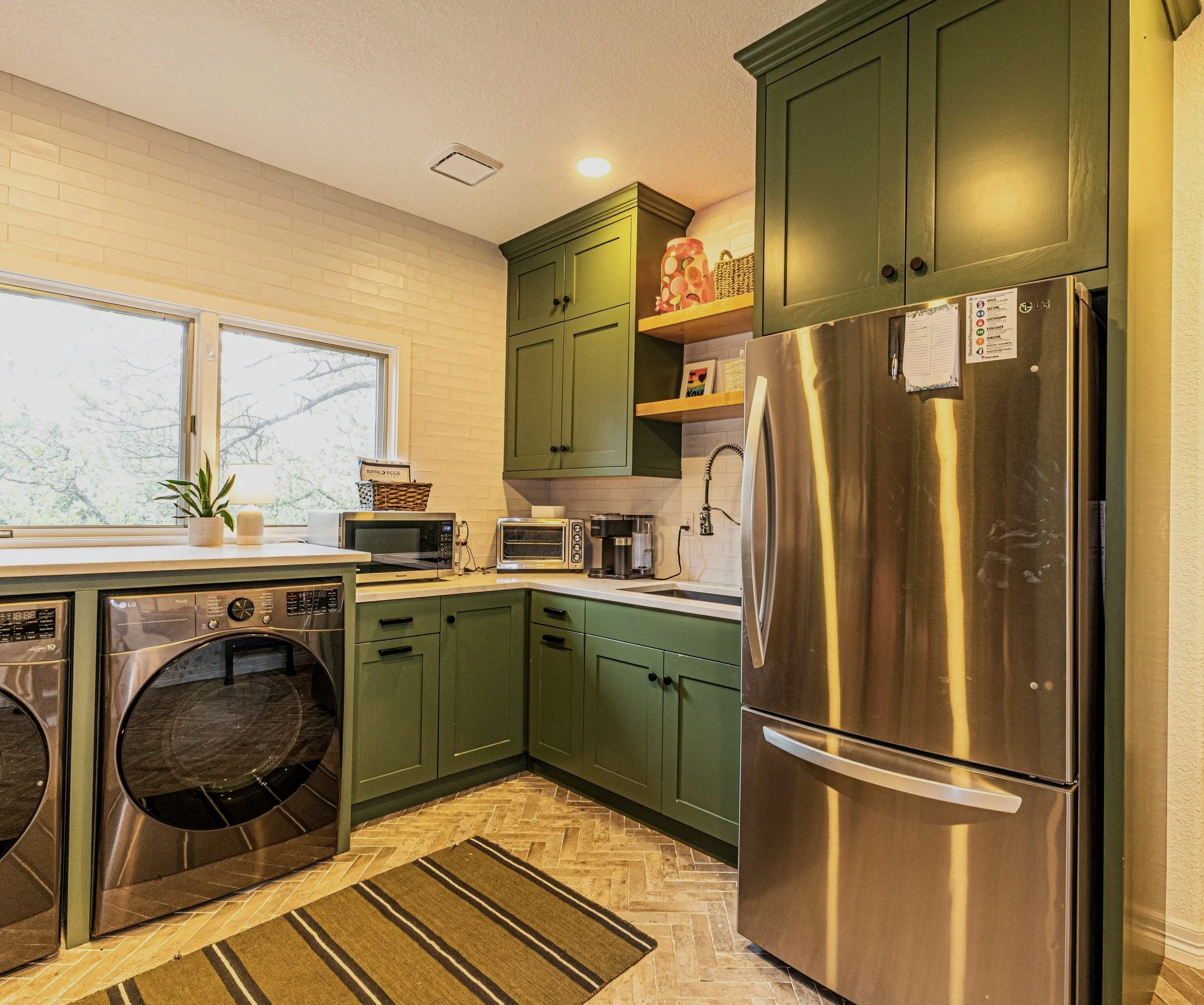 Kitchen with green cabinets, stainless steel refrigerator, black and stainless steel appliances, window with a view outside, plants, and a striped rug on a brown patterned floor.