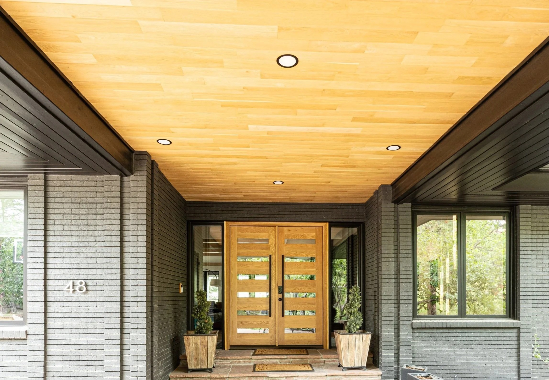 View of a home's entryway with a modern wooden door, flanked by two large planters and windows, in front of gray brick walls and a ceiling with recessed lighting.