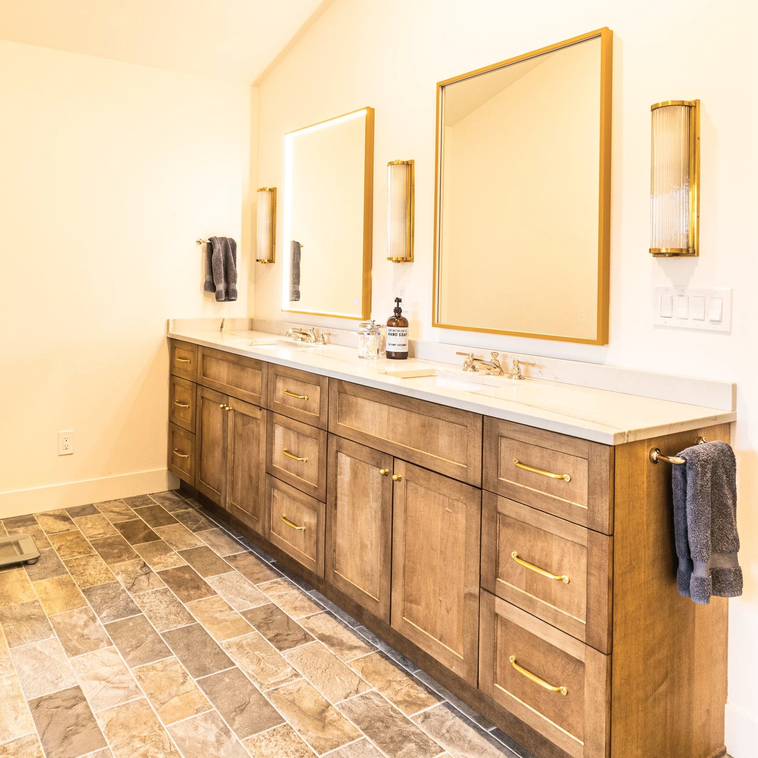 Bathroom vanity with two large mirrors, gold frames, and gold fixtures. Beige towel hanging on a towel rack, soap and hand sanitizer bottles on the counter, granite countertops, and a brown wooden cabinet with drawers.