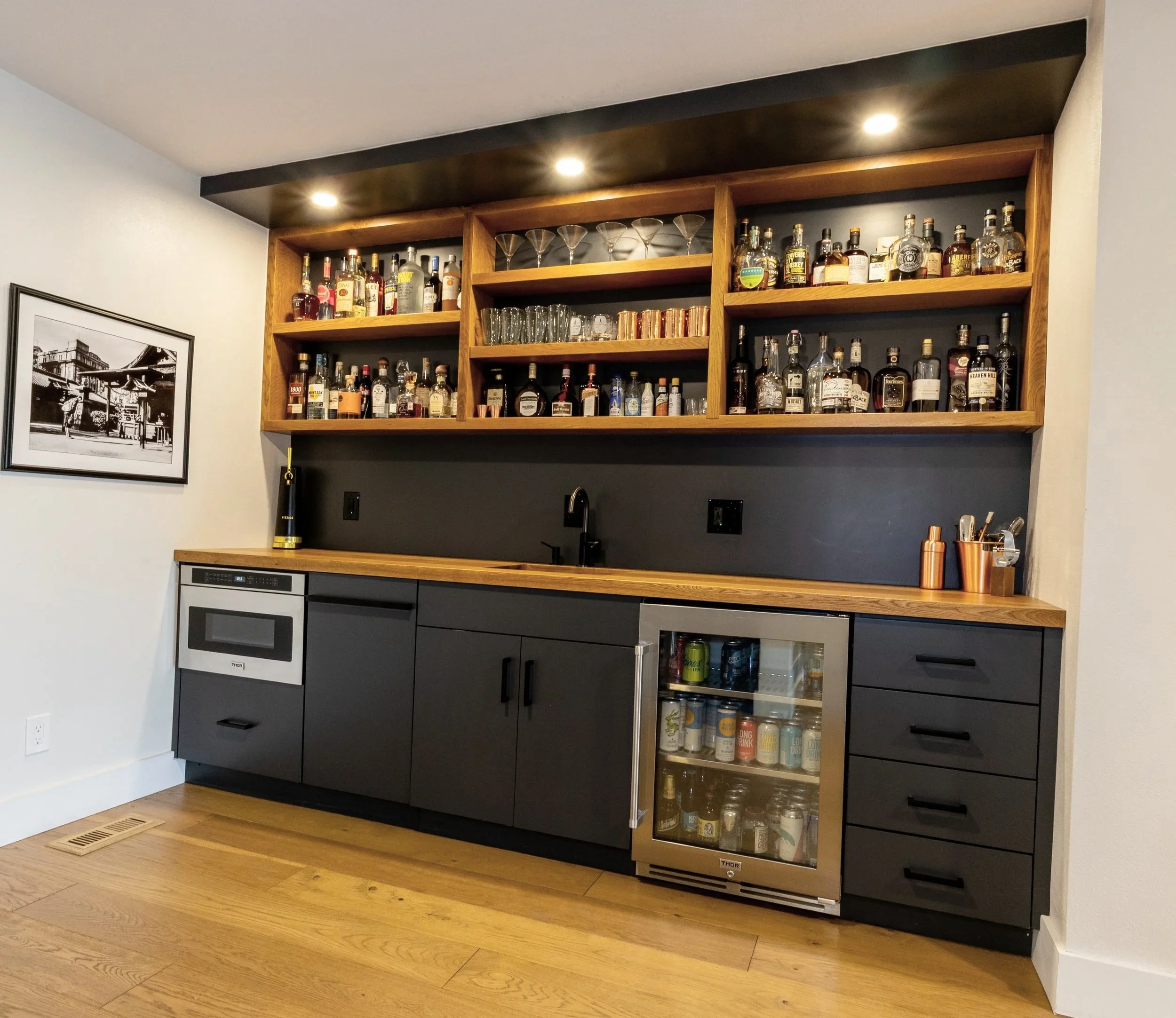 A modern kitchen bar area with wooden and black cabinetry, featuring a built-in wine cooler, and open shelves displaying liquor bottles, glasses, and cans. Recessed ceiling lights illuminate the space; a black and white framed photo hangs on the wall