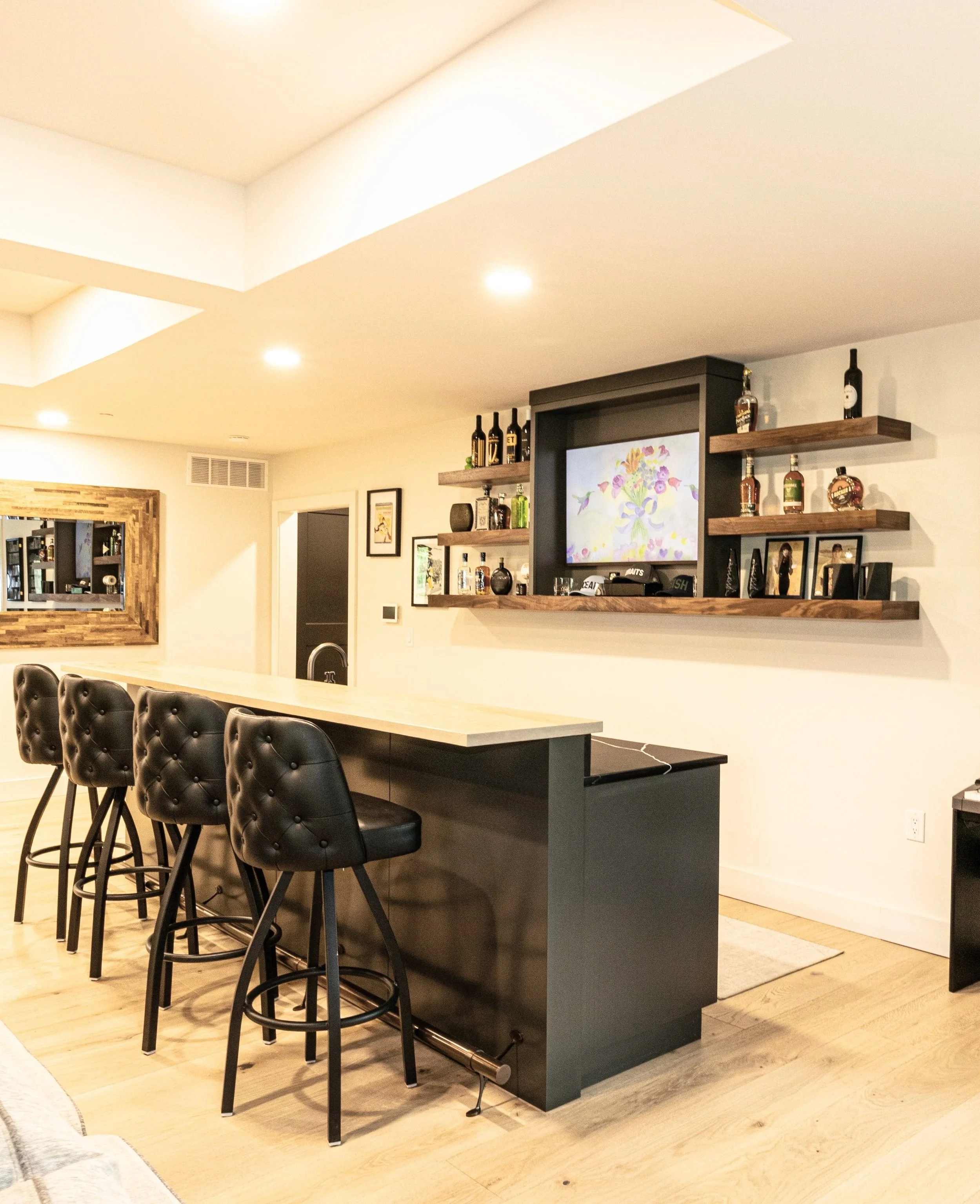 Interior view of a modern kitchen or bar area with a high counter, black leather chairs, and a shelf with bottles of alcohol and framed photos. A large flat-screen TV displays a flower graphic.