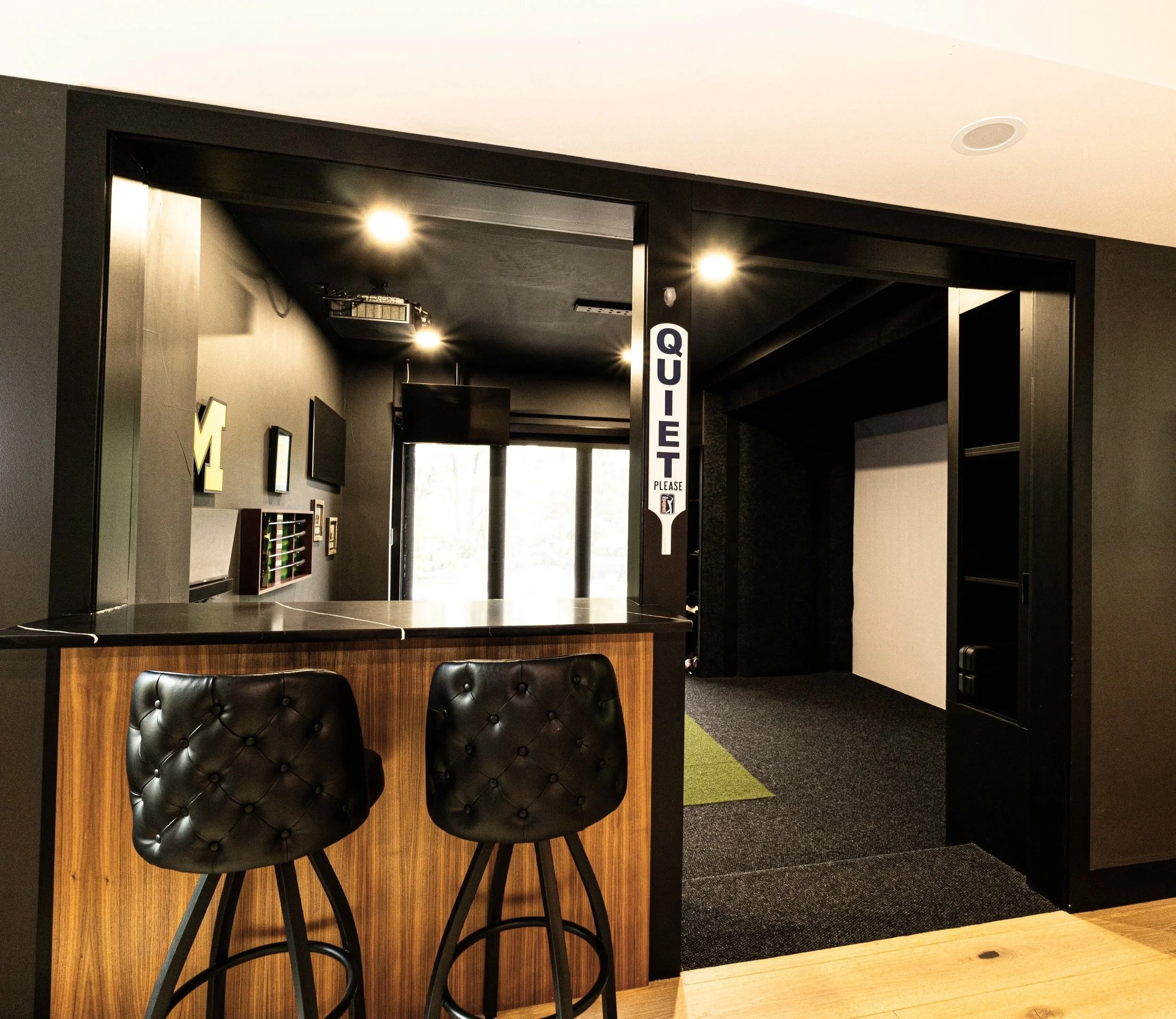 Interior view of a modern office or lobby area with black chairs at a wooden counter, dark walls, and a ceiling with spotlights, adjacent to a corridor with a sign labeled 'QUALITY PLEASE'.
