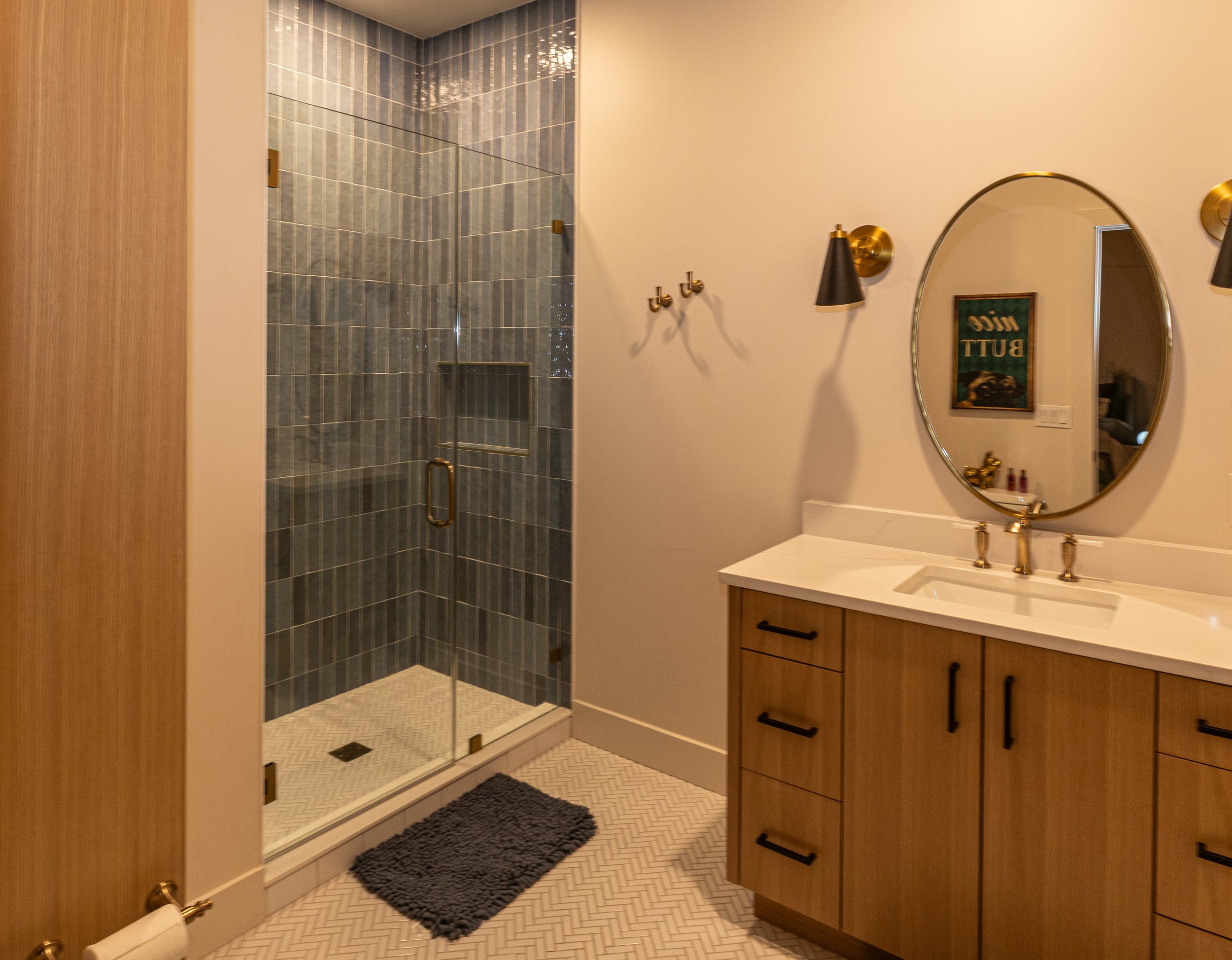 Bathroom featuring a glass-enclosed walk-in shower with blue tiles, a black and gold wall sconce, oval mirror, and wooden vanity with black handles.